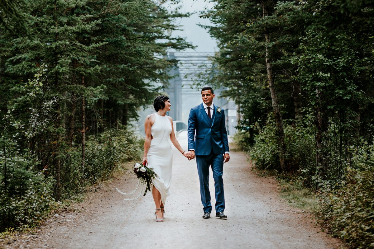 A couple in wedding attire walks hand in hand down a forest path near Canmore, surrounded by lush green trees. The bride holds a bouquet and wears a white dress, while the groom is in a blue suit. They look at each other and smile.