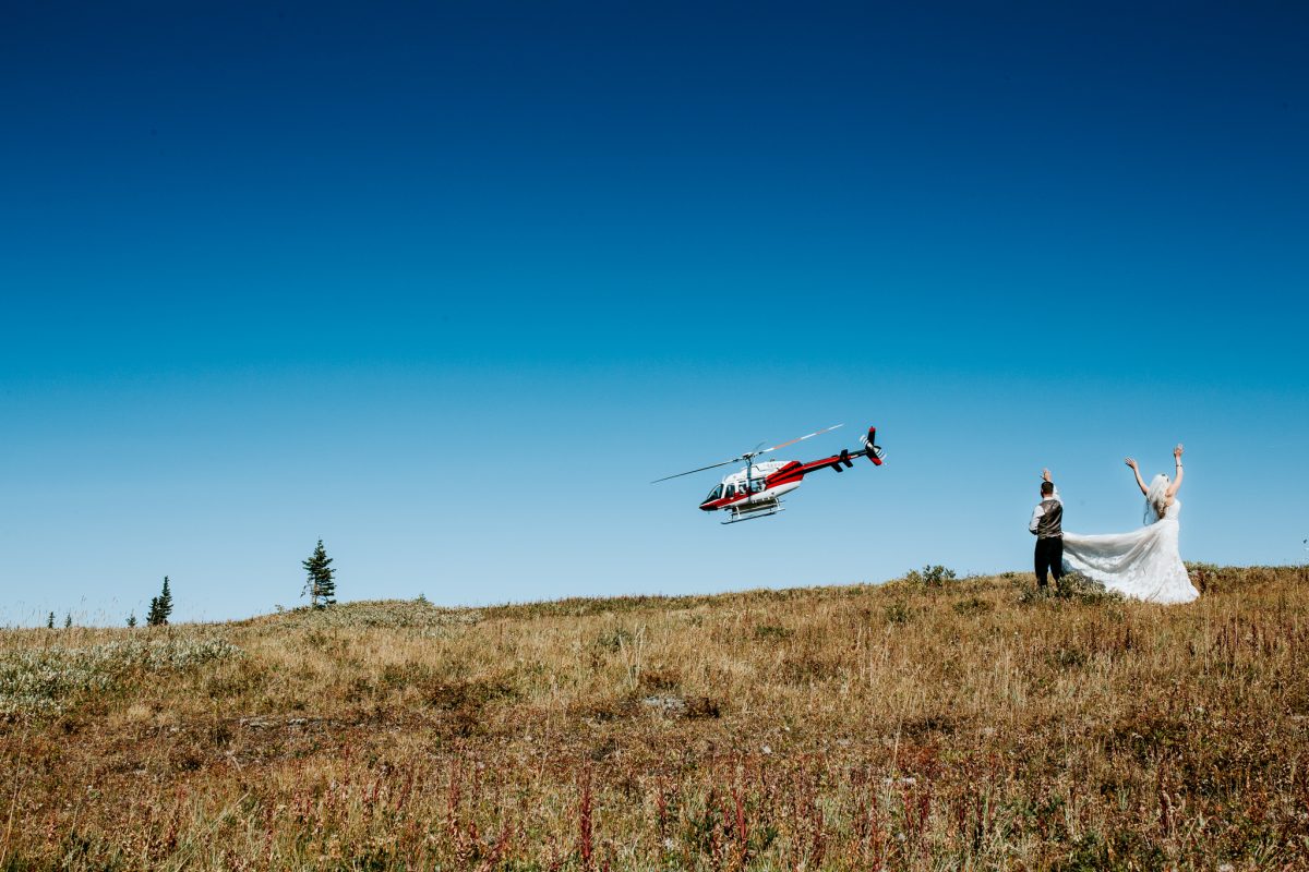 A bride and groom stand on a grassy hill, waving as a red and white helicopter flies nearby under a clear blue sky—an unforgettable elopement moment captured by their photographer.