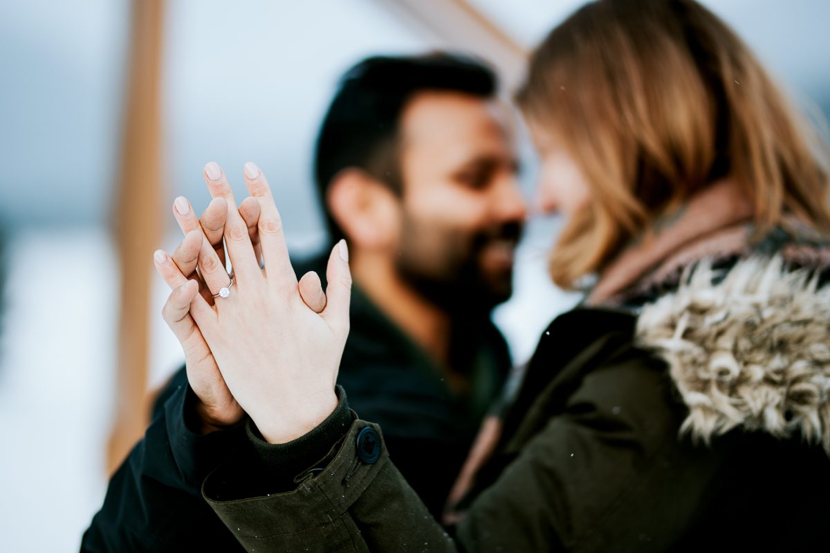 A close-up of a couple holding hands outdoors in Banff, with the focus on a woman's hand showing an engagement ring. Both are smiling and their faces are softly blurred, beautifully captured by a skilled photographer.