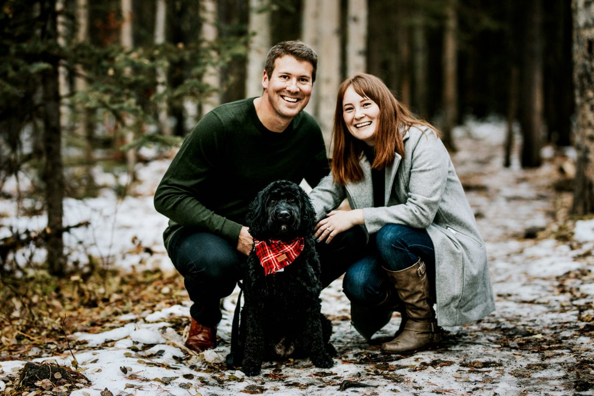 A smiling man and woman kneel next to a black curly-haired dog wearing a red plaid bandana in a snowy Banff forest, surrounded by trees and fallen leaves.