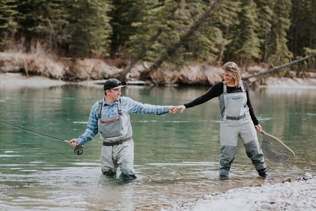 A man and woman in fishing gear hold hands and smile while standing in a river surrounded by trees near Canmore; the man holds a fishing rod and the woman holds a net.