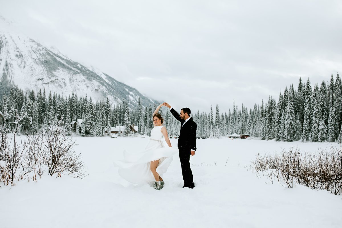 A bride and groom dance in the snow near Canmore, surrounded by snow-covered trees and mountains, with cozy cabins in the background on a cloudy winter day—perfect for an elopement photographer to capture unforgettable moments.