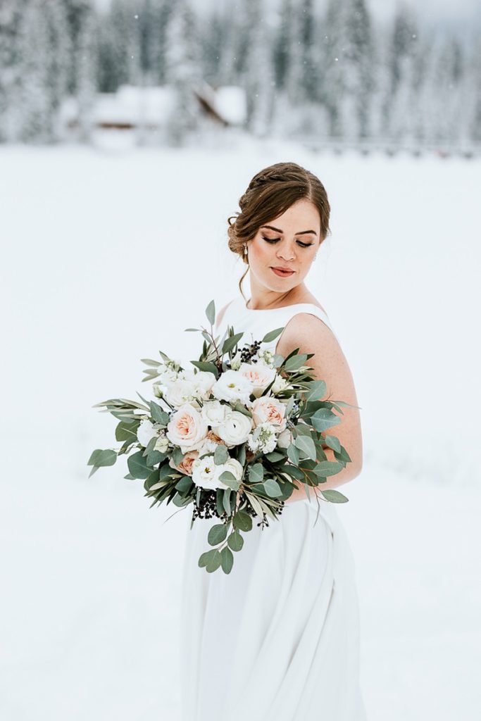 A bride in a white dress stands in a snowy Calgary landscape, holding a large bouquet of white and blush flowers with greenery. Captured by a talented wedding photographer, she looks down, with trees and a house blurred in the background.