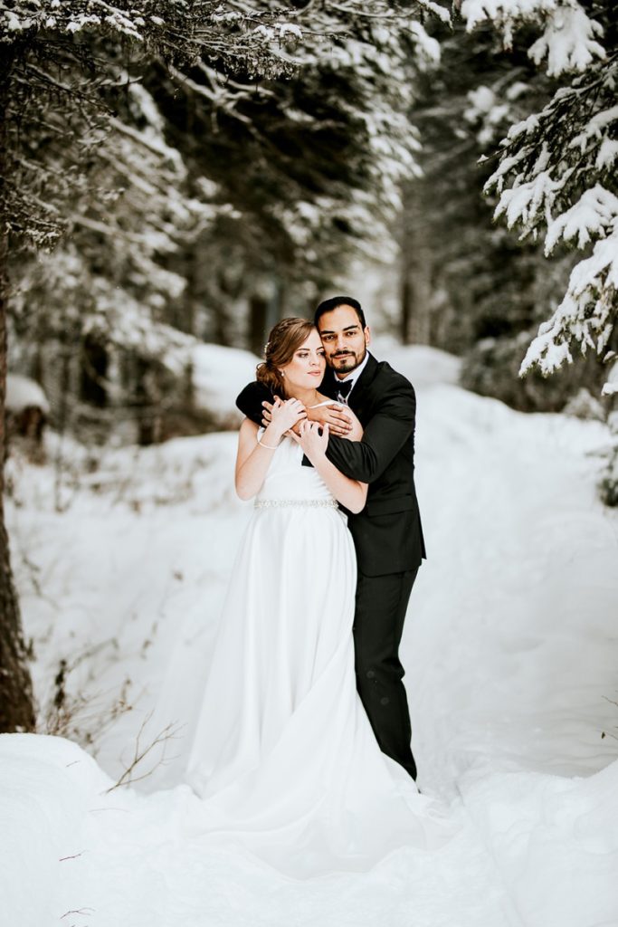 A bride and groom stand together on a snowy path surrounded by snow-covered trees in Canmore. The groom embraces the bride from behind, both smiling and dressed in formal wedding attire, capturing a magical winter elopement.