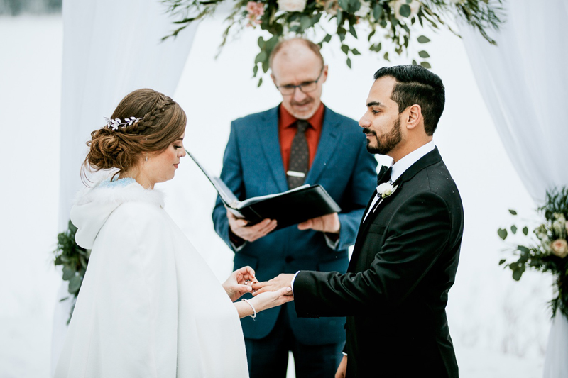 A bride and groom exchange rings under a floral arch during a winter outdoor ceremony in Banff, with an officiant standing behind them. Captured by a skilled photographer, the bride wears a white cloak and the groom a black suit.
