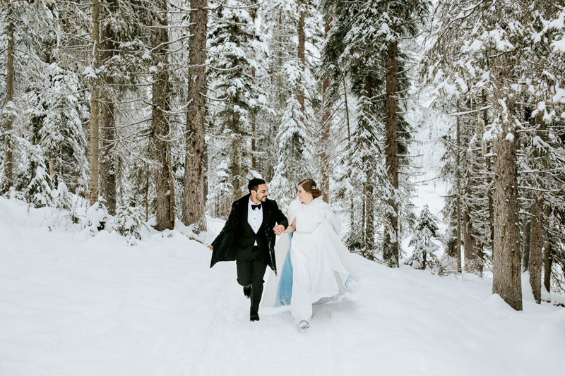 A couple in formal wedding attire walks hand in hand through a snowy Banff forest, surrounded by tall, snow-covered trees. The scene looks joyful and wintry.