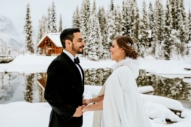 A bride and groom stand outdoors in the snow, holding hands and smiling at each other. Behind them are snowy trees and a wooden cabin by a reflective lake, capturing a magical Banff elopement winter wedding scene.