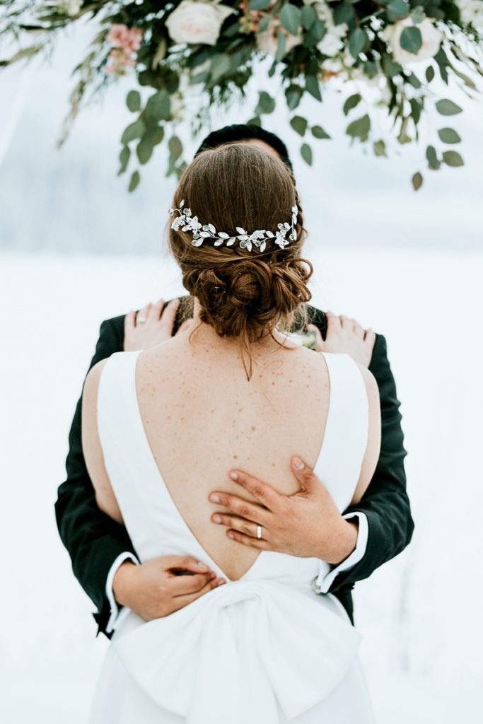 A bride in a white dress with a jeweled hairpiece embraces her groom in a dark suit in a snowy outdoor setting near Calgary, standing under greenery and flowers. Captured by their wedding photographer, the groom’s ringed hand rests on her back.