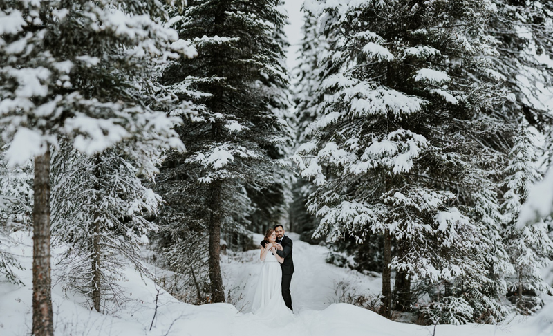 A couple in wedding attire stands closely together in a snowy forest near Calgary, surrounded by tall pine trees covered in snow. The scene is serene and romantic, beautifully captured by a talented elopement photographer.