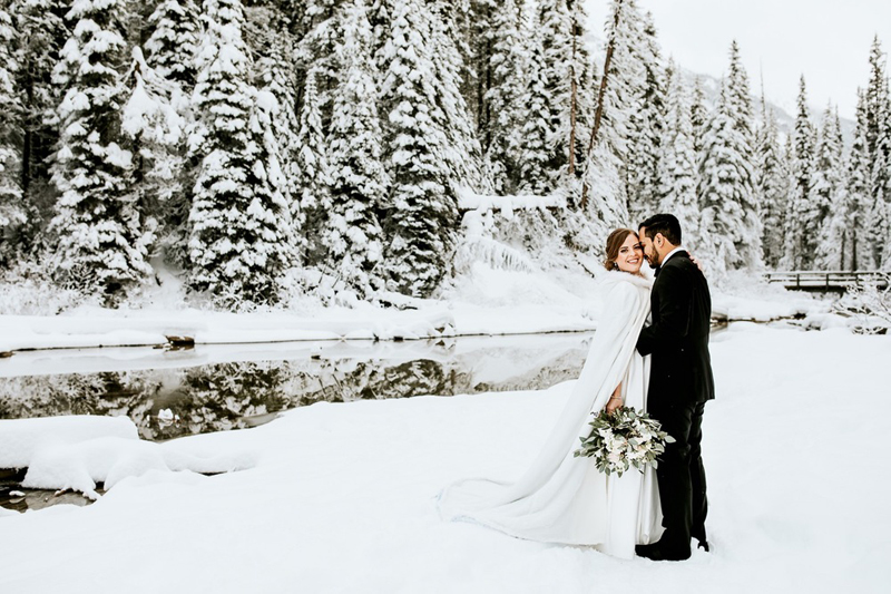 A bride and groom stand together in a snowy Banff forest beside a river, with tall snow-covered trees in the background. The bride wears a white cape and holds a bouquet, while the groom wears a black suit—perfect for an elopement photographer to capture.