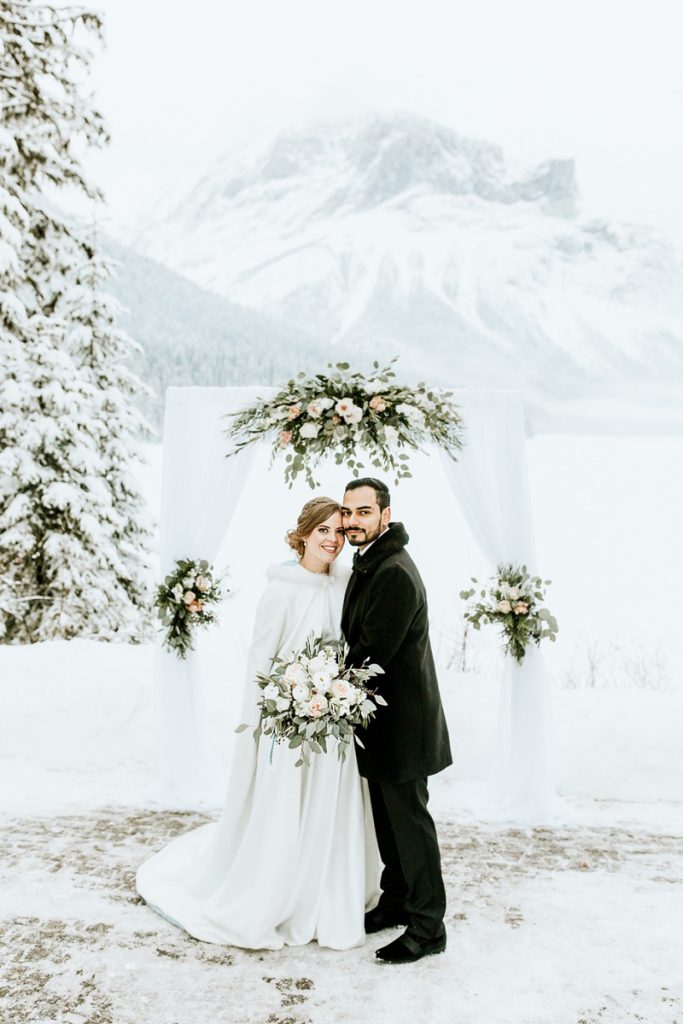 A bride and groom stand smiling in front of a white floral wedding arch outdoors in snowy Canmore, with evergreen trees and a snow-covered mountain in the background, beautifully captured by their photographer.