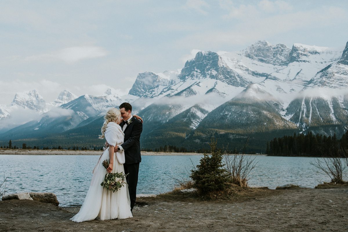 A bride and groom embrace near a lake with snow-capped mountains in the background, surrounded by pine trees and natural scenery under a partly cloudy sky—a perfect Canmore elopement.