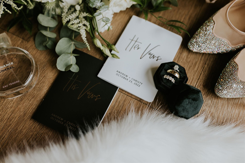 Close-up of wedding details on a wooden surface: Calgary photographer captures two His Vows booklets, a white flower bouquet, sparkling shoes, rings in a green box, perfume bottle, and white fur fabric in the foreground.