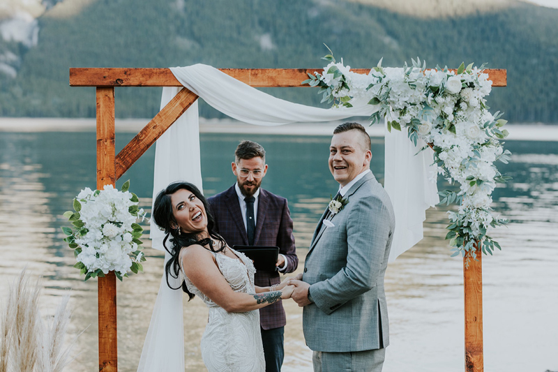 A smiling couple stands under a wooden wedding arch decorated with white flowers and fabric by a lake near Banff, holding hands as an officiant in a suit stands behind them. Mountains and trees are visible in the background.