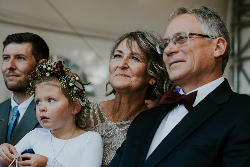A group of four people, including a young girl with a flower crown, an older woman, and two men in formal attire, stand close together and look ahead, smiling warmly for a Calgary photographer.
