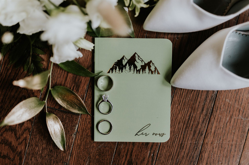 A green wedding notebook with “her vows” and a mountain design rests on a wooden floor, beside white flowers, white high-heeled shoes, and three rings—including an engagement ring—perfect for capturing by any photographer.