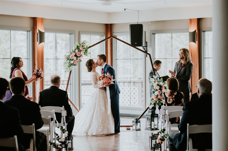 A bride and groom share a kiss at their indoor wedding ceremony in Canmore, standing under a geometric floral arch, surrounded by seated guests and their photographer capturing the moment.