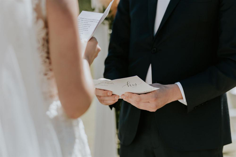 A bride and groom stand facing each other, holding handwritten vows during their Banff wedding ceremony. The bride wears a white dress, and the groom is in a black suit. Their faces are not visible.
