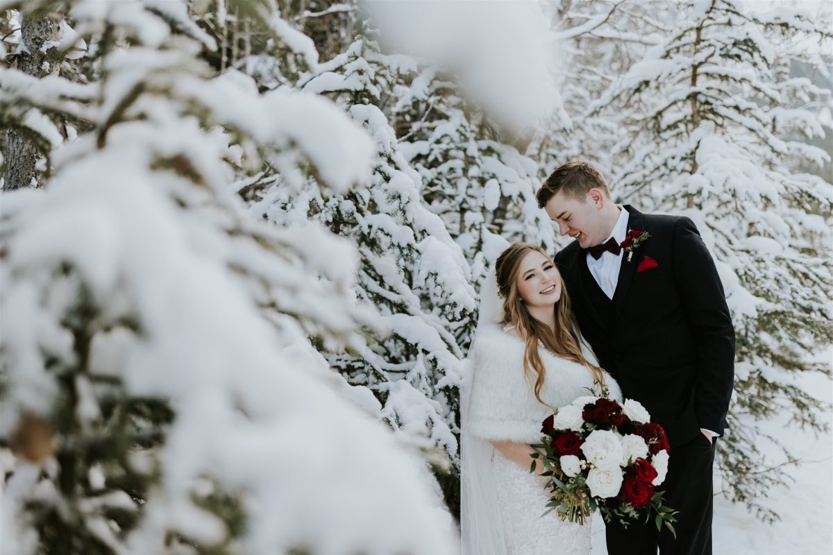 A bride and groom stand together in a snowy forest near Canmore. The bride holds a bouquet of red and white flowers and wears a white fur wrap, while the groom, in a black suit with a bow tie, leans his head toward her as their photographer captures the moment.