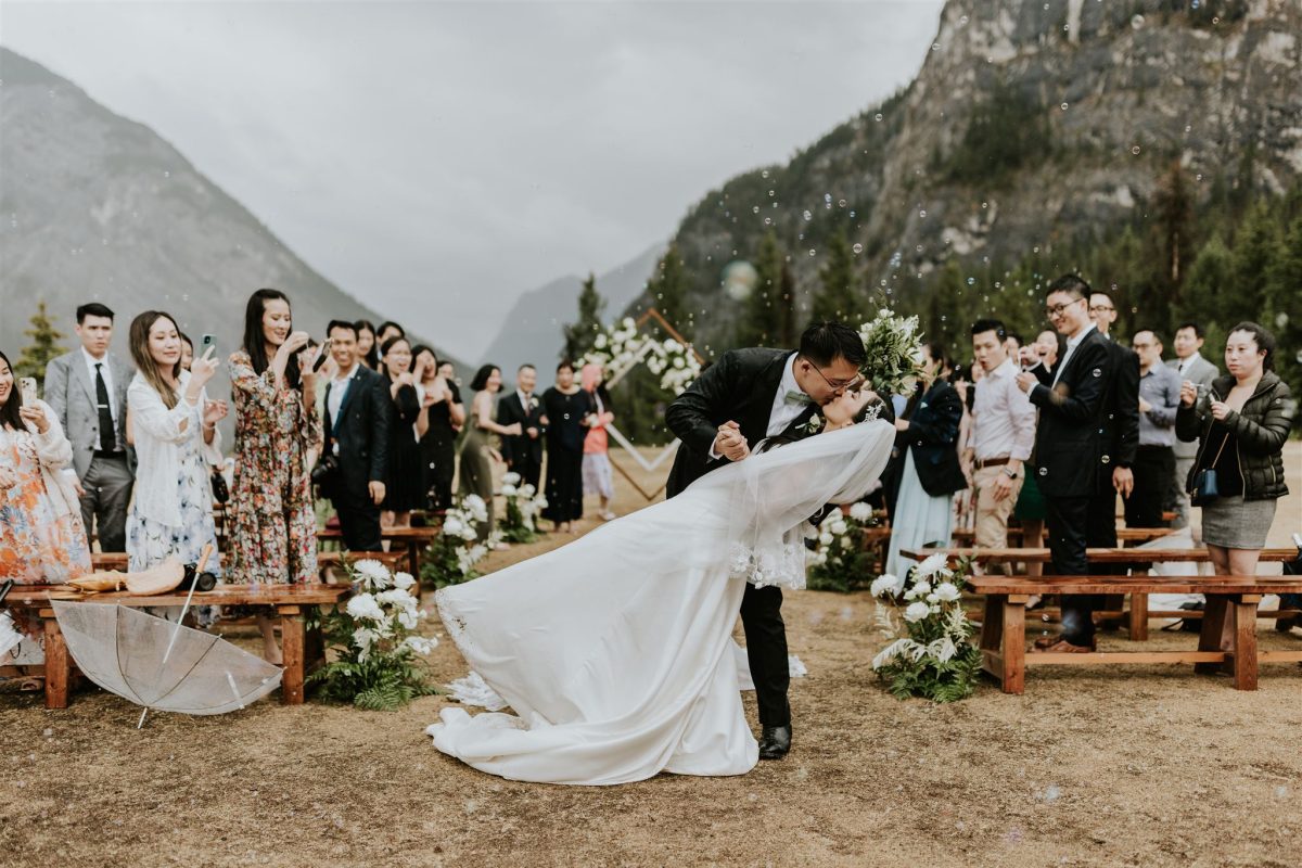 A bride and groom share a dramatic kiss outdoors after their wedding ceremony in Canmore, surrounded by cheering guests, with mountains and floral arrangements in the background—perfect for any photographer to capture.