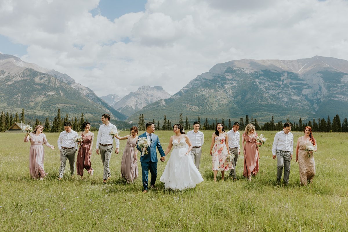 A bride and groom walk hand in hand with their wedding party in a grassy field, surrounded by friends in pastel dresses and shirts, as a Banff photographer captures their special elopement amid mountains and cloudy skies.