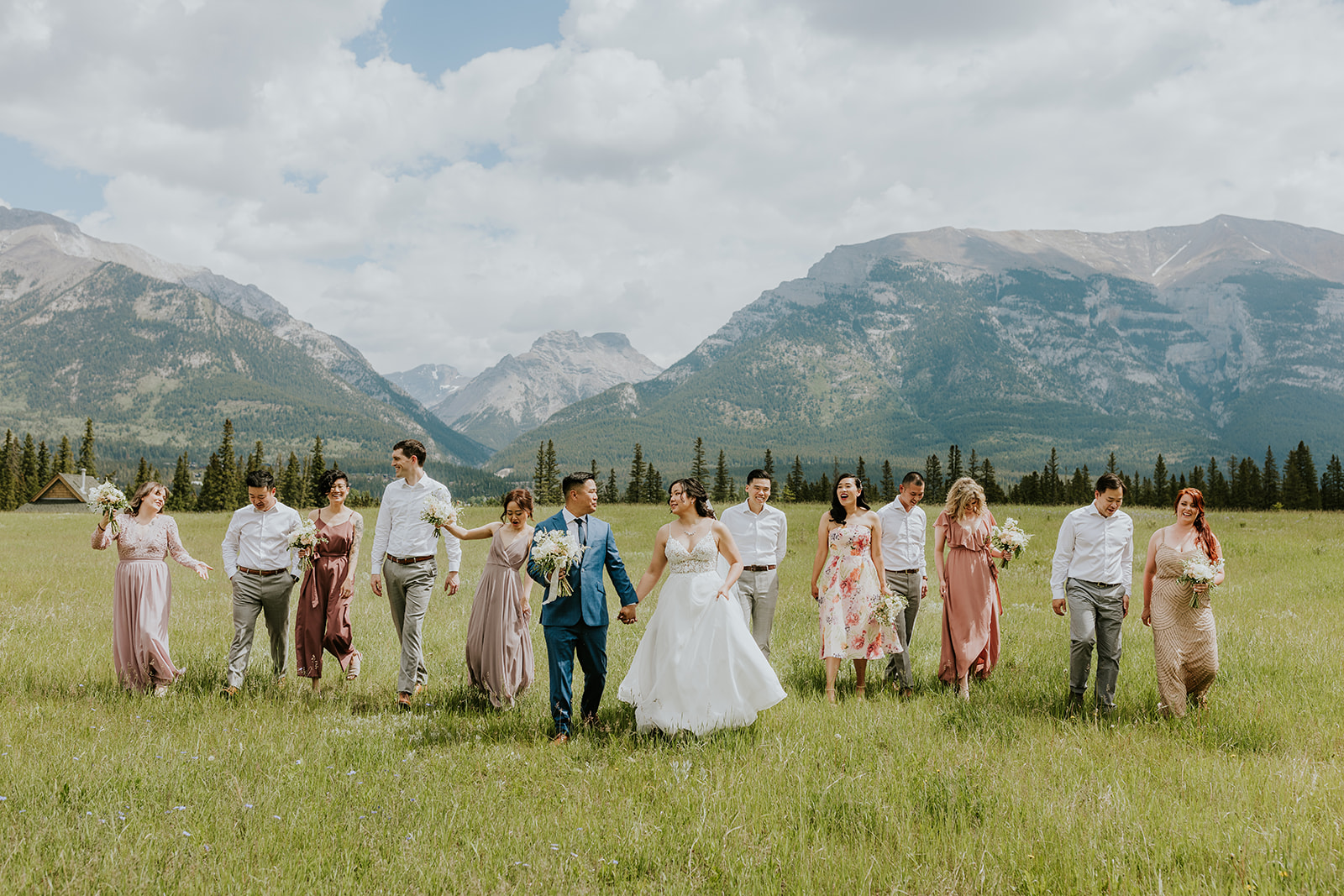 A bride and groom walk hand in hand with their wedding party in a grassy field, surrounded by friends in pastel dresses and shirts, as a Banff photographer captures their special elopement amid mountains and cloudy skies.