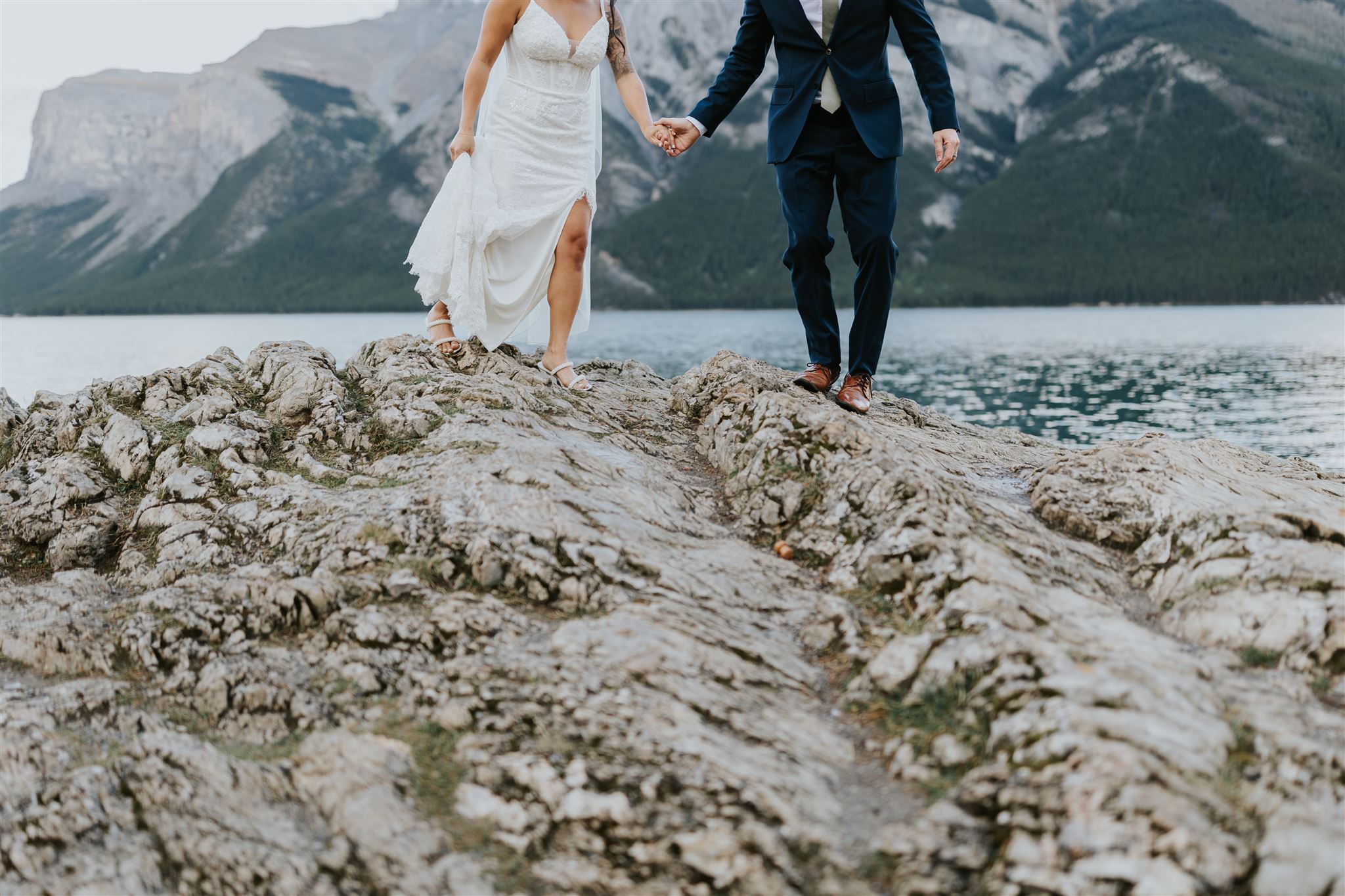 A bride in a white dress and a groom in a navy suit walk hand in hand on rocky terrain by a lake, with Banff’s mountains and trees in the background—an elopement beautifully captured by their photographer. Their faces are out of frame.