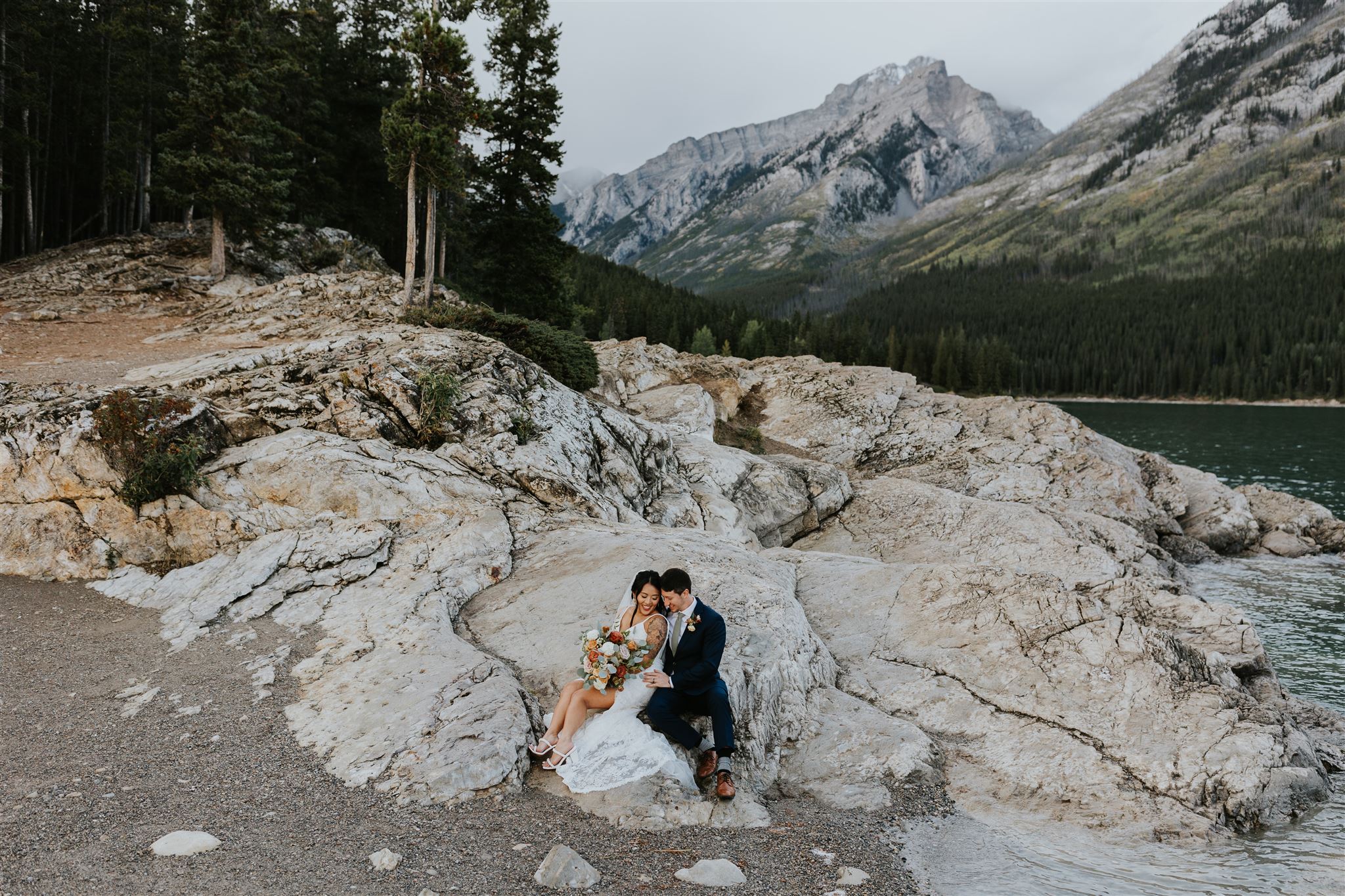 A bride and groom sit together on rocky terrain by a lake near Banff, surrounded by pine trees and mountains, sharing an intimate moment in nature captured beautifully by their photographer.