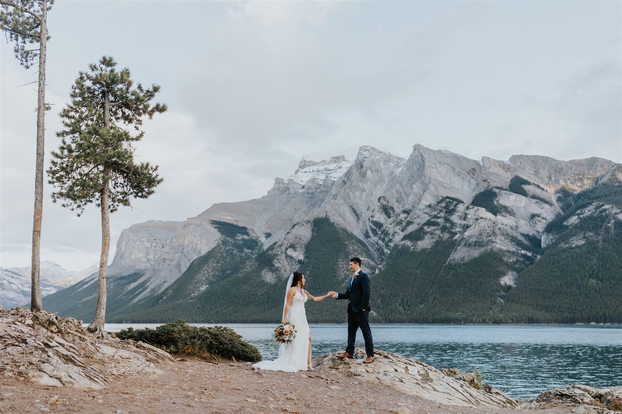A bride and groom stand on a rocky lakeshore near Canmore, holding hands with a bouquet at their feet. Behind them are pine trees, a calm lake, and towering mountains under a cloudy sky—captured beautifully by their photographer.