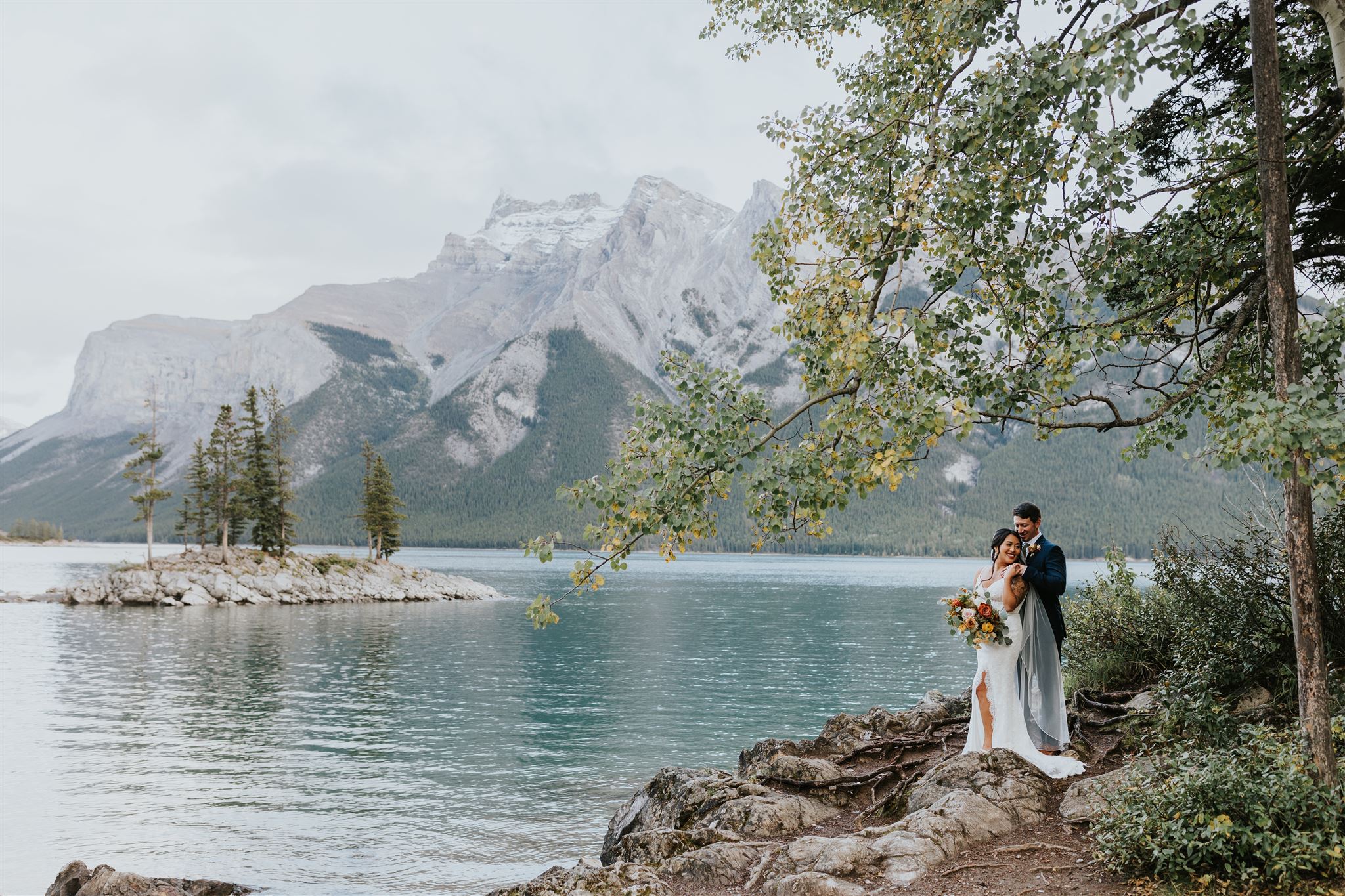 A bride and groom stand on rocky ground by a calm lake in Canmore, embracing under a tree. Mountains and a small island with pine trees are visible in the background, creating a serene, scenic wedding landscape.