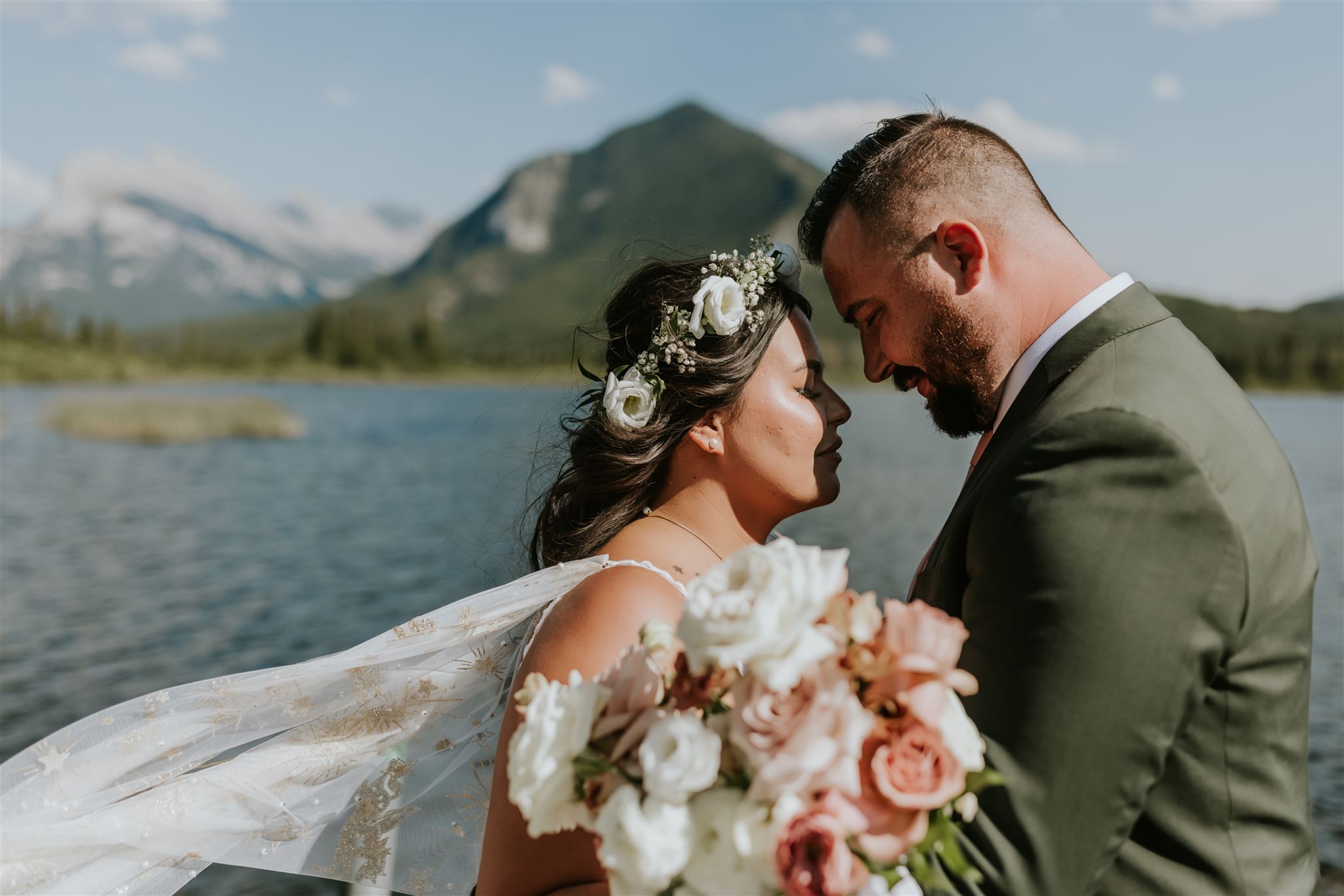 A bride and groom embrace by a lake with mountains in the background near Canmore. The bride wears a floral crown and veil, holding a bouquet as they gently touch foreheads under a blue sky—perfect for an intimate elopement.