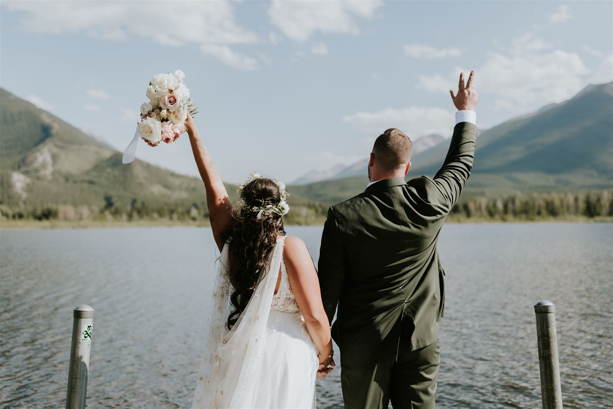A bride and groom stand hand in hand on a dock in Canmore, facing a lake and mountains. The bride lifts her bouquet while the groom celebrates, all beautifully captured by their elopement photographer under a partly cloudy sky.