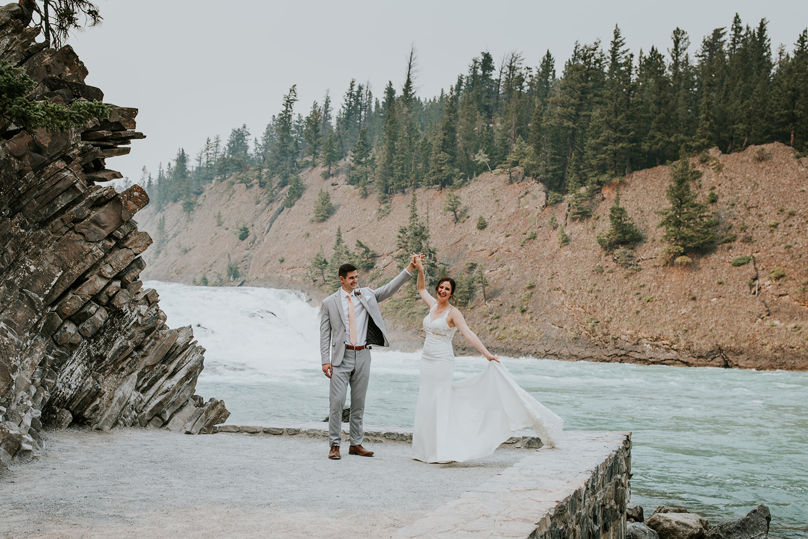 A bride and groom pose by a river with rocky cliffs and pine trees in the background. Captured by their Calgary photographer, the groom holds the bride’s hand as she twirls, both smiling joyfully during their elopement outdoors.