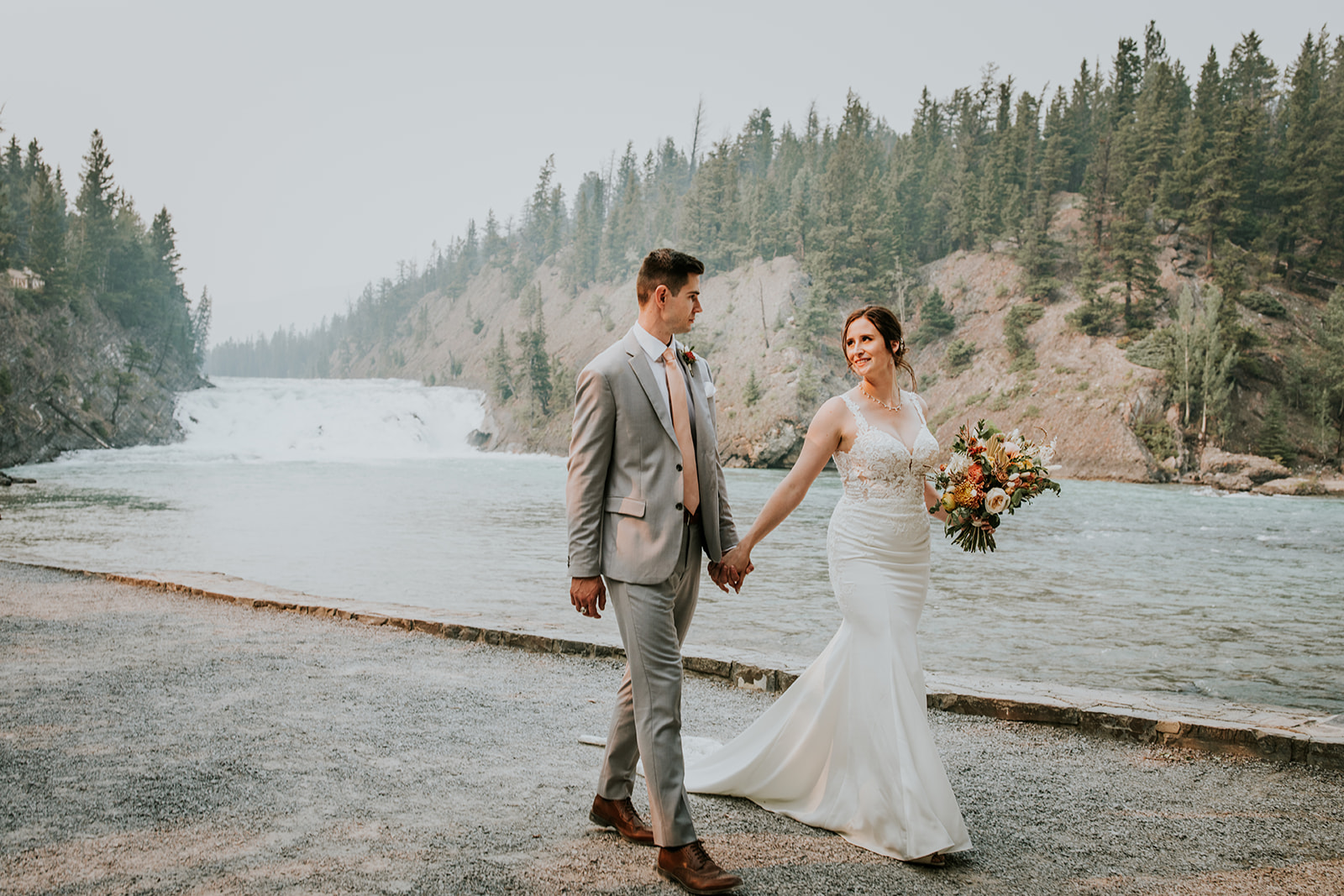 A bride in a white dress and a groom in a light gray suit hold hands and walk by a river in Banff, with a waterfall and pine trees behind them. The bride carries a colorful bouquet, smiling at her partner during their elopement.