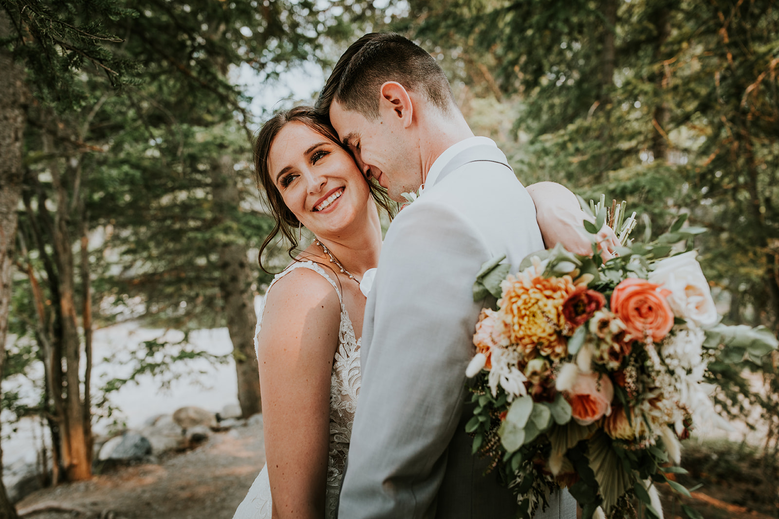 A bride and groom embrace outdoors among trees in Canmore. The bride smiles at the camera with a colorful bouquet, while the groom gently rests his head on her temple. Both wear wedding attire, capturing a beautiful elopement moment.