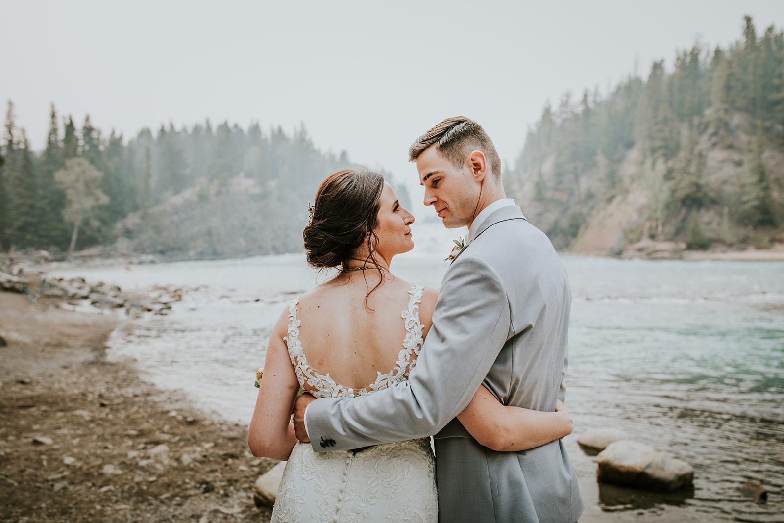 A bride and groom embrace and gaze at each other by a river, surrounded by trees and rocky shoreline, on a misty day—capturing the essence of an intimate wedding for their photographer.