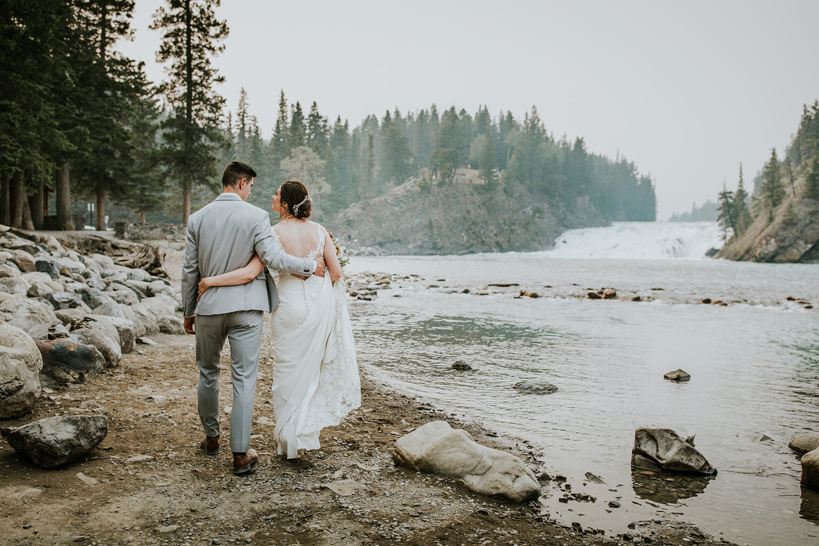 A bride and groom walk arm in arm along a rocky riverbank in Banff, with tall trees and a waterfall in the background under a cloudy sky—perfect for a wedding photographer to capture unforgettable moments.