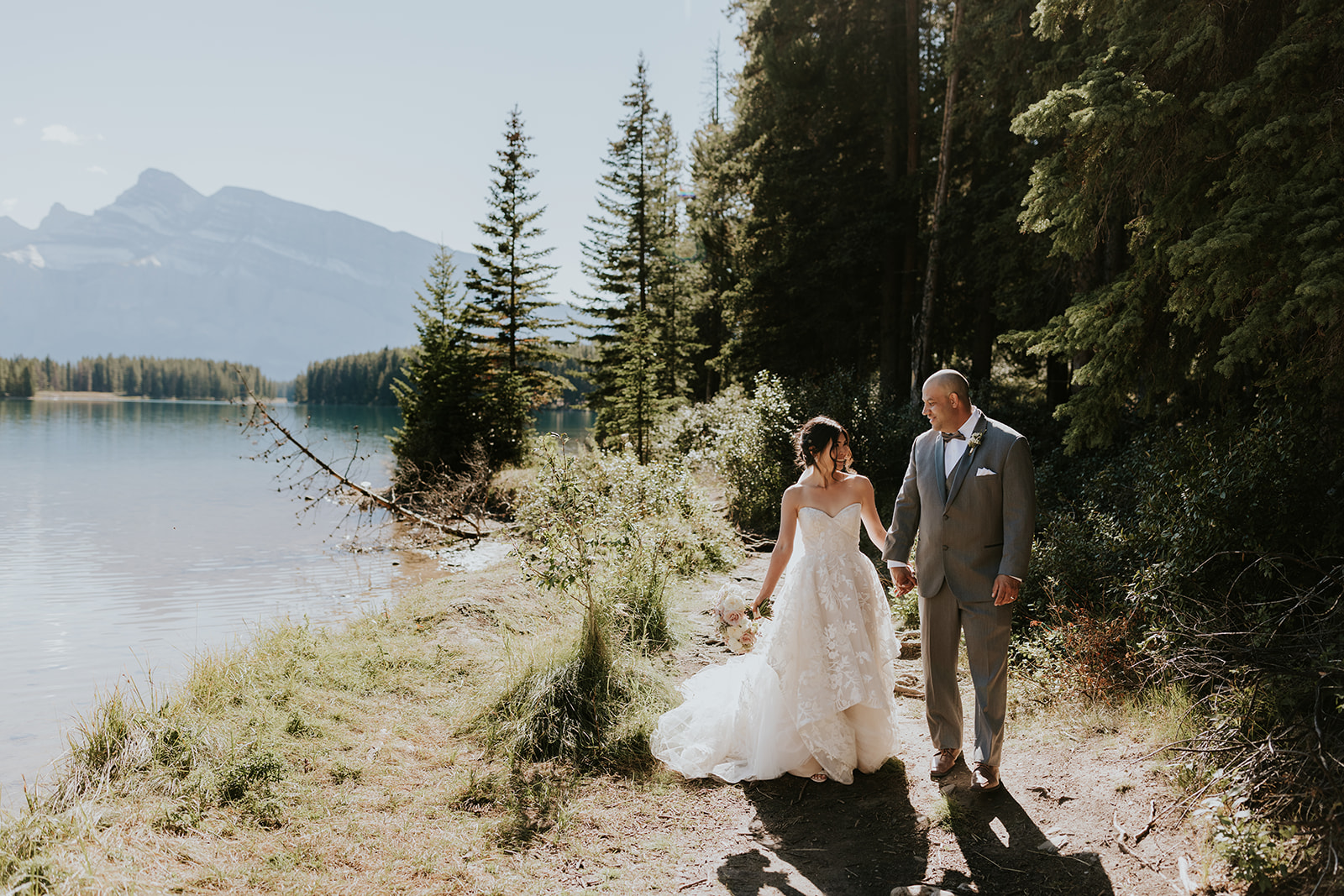 A bride in a white gown and a groom in a gray suit walk hand in hand along a forested lakeshore, as their Calgary elopement photographer captures pine trees and distant mountains beneath a clear sky.