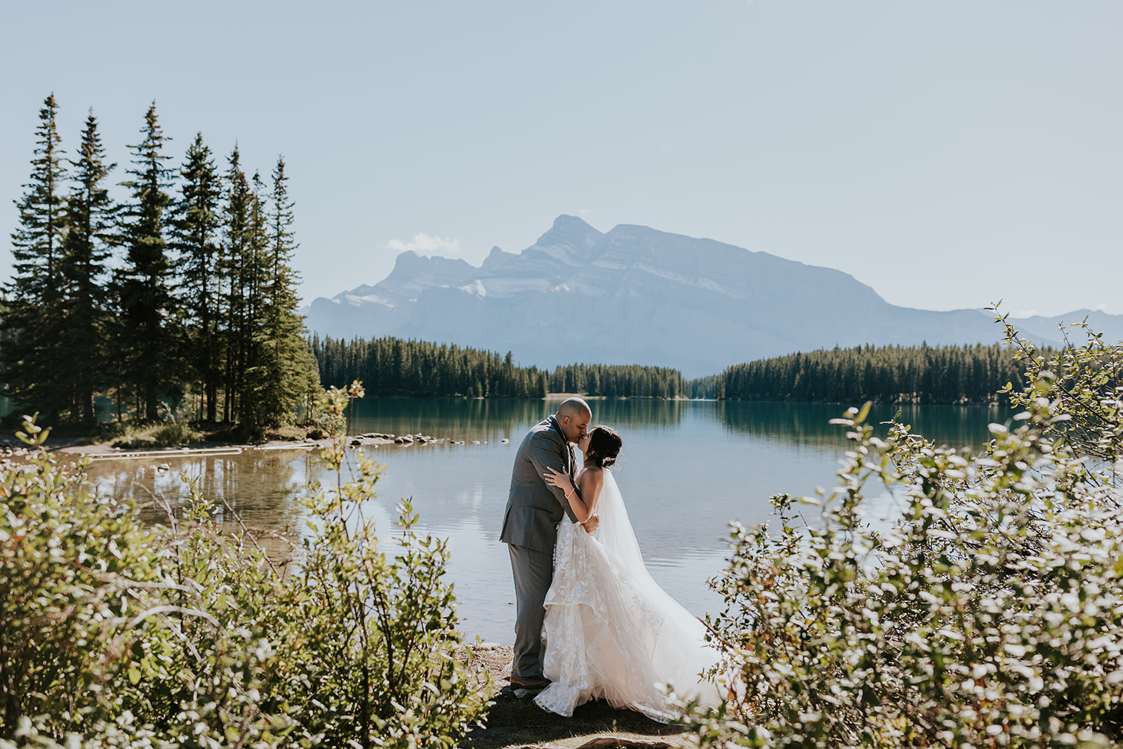 A bride and groom embrace by a serene lake in Canmore, surrounded by greenery and pine trees, with tall mountains and a clear blue sky—capturing the perfect elopement moment.