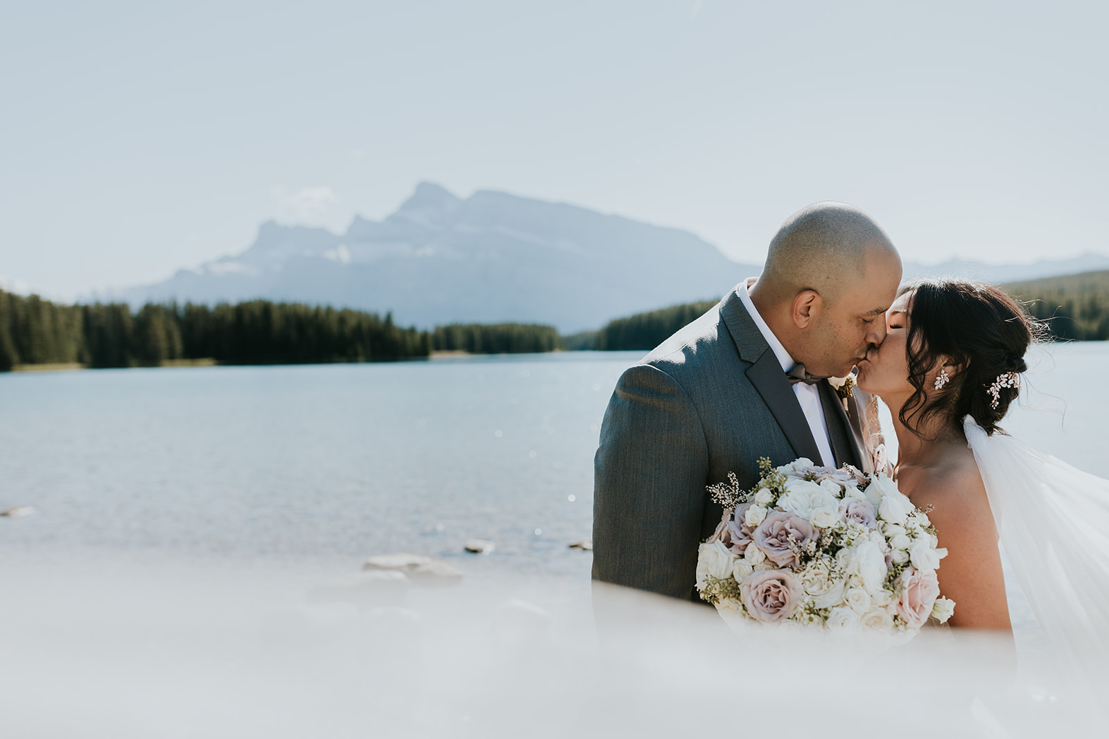 A bride and groom share a kiss by a serene lake with mountains in the background, celebrating their Calgary elopement. The bride holds a bouquet of white and pale pink flowers, both dressed in wedding attire under clear blue skies.