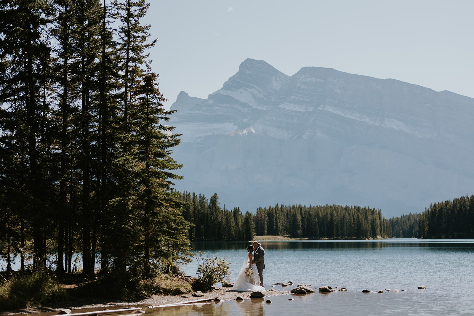 A couple in wedding attire stands on rocks by a lake in Banff, surrounded by trees, with a large mountain and clear sky in the background, captured beautifully by their photographer.