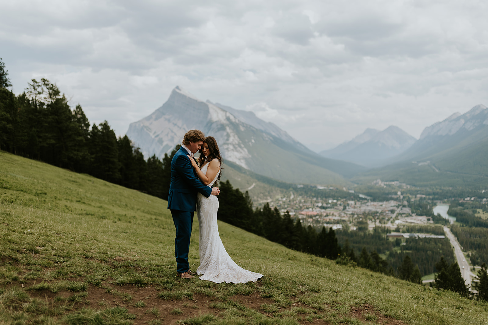 A bride and groom embrace on a grassy hillside as a photographer captures their love, with forest and a mountain valley near Canmore in the background under a cloudy sky.