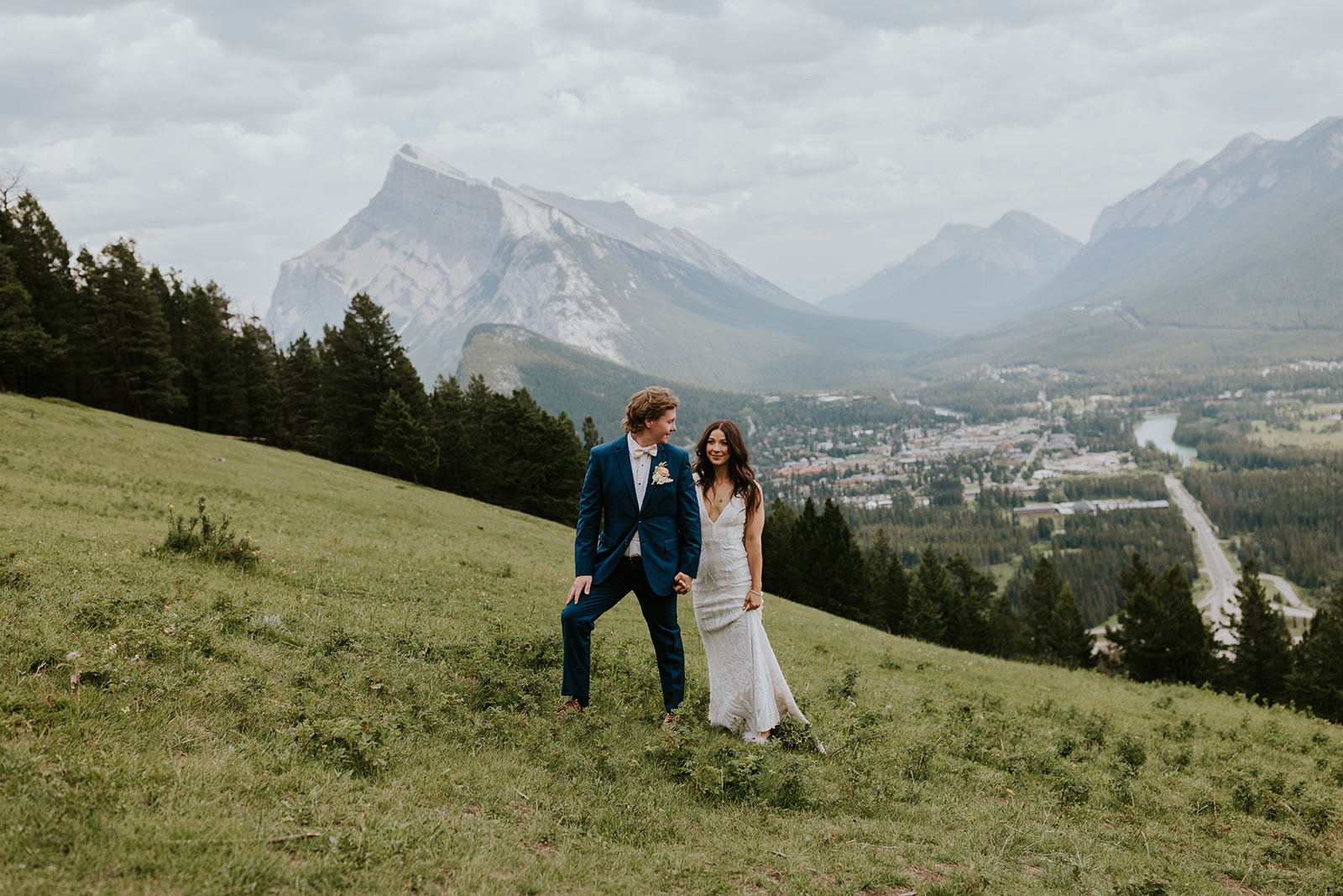 A bride and groom smile and hold hands while walking on a grassy hillside near Canmore, with mountains, forests, and a valley town in the background on a cloudy day.