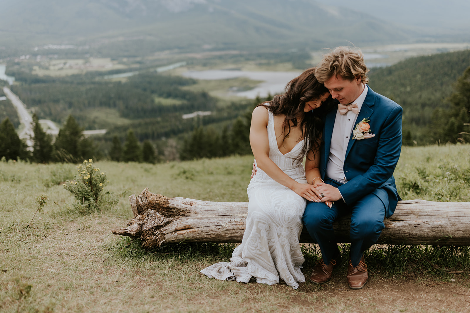 A bride and groom sit on a log outdoors near Canmore, heads touching and holding hands, framed by mountains, forests, and a winding river. The bride wears a white dress while the groom sports a stylish blue suit.