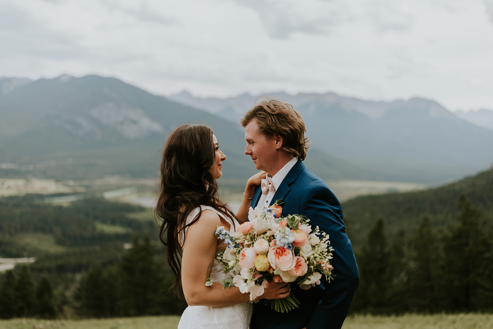 A bride and groom stand close together, gazing at each other and smiling, with the bride holding a colorful bouquet. They are outdoors at a Canmore wedding, surrounded by mountains and forest in the background.