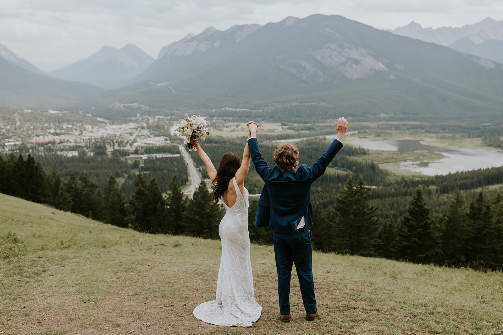 A bride in a white dress and a groom in a blue suit stand on a grassy hill with arms raised, overlooking a scenic Banff valley, forests, and distant mountains under a cloudy sky—captured perfectly by their wedding photographer.