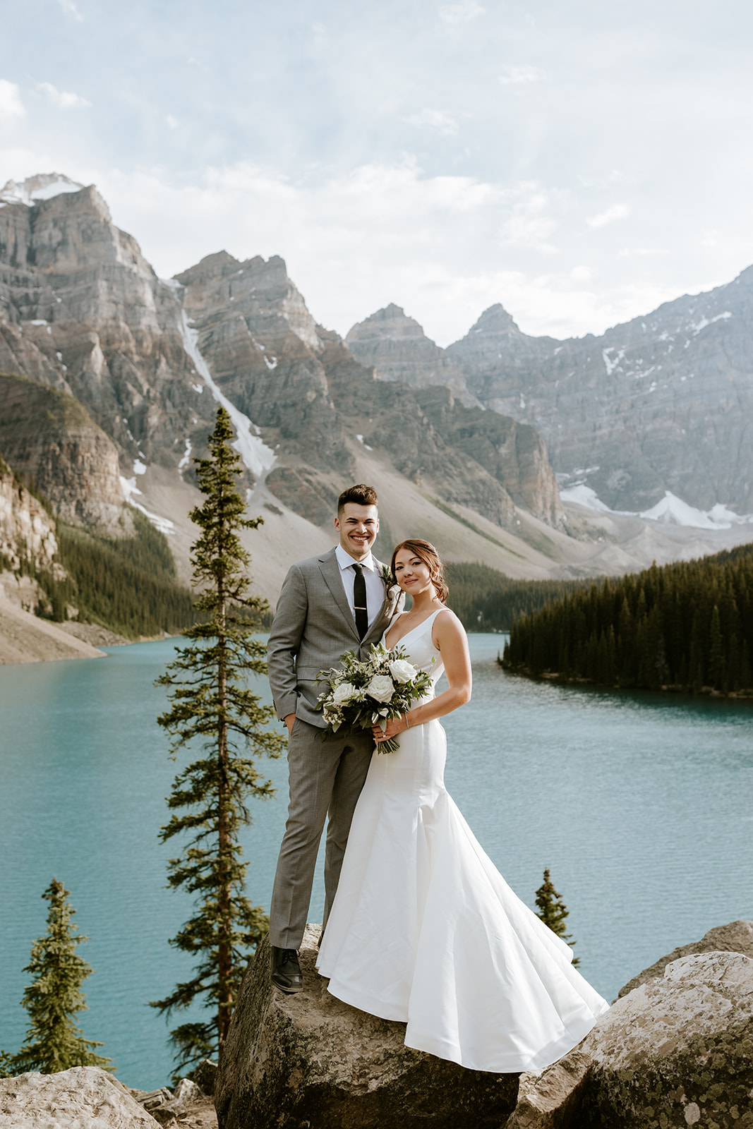 A bride and groom stand on a rock by a turquoise lake, surrounded by tall pines and mountains. Captured by a Calgary elopement photographer, the scene is bright and serene as the couple celebrates in nature's beauty.