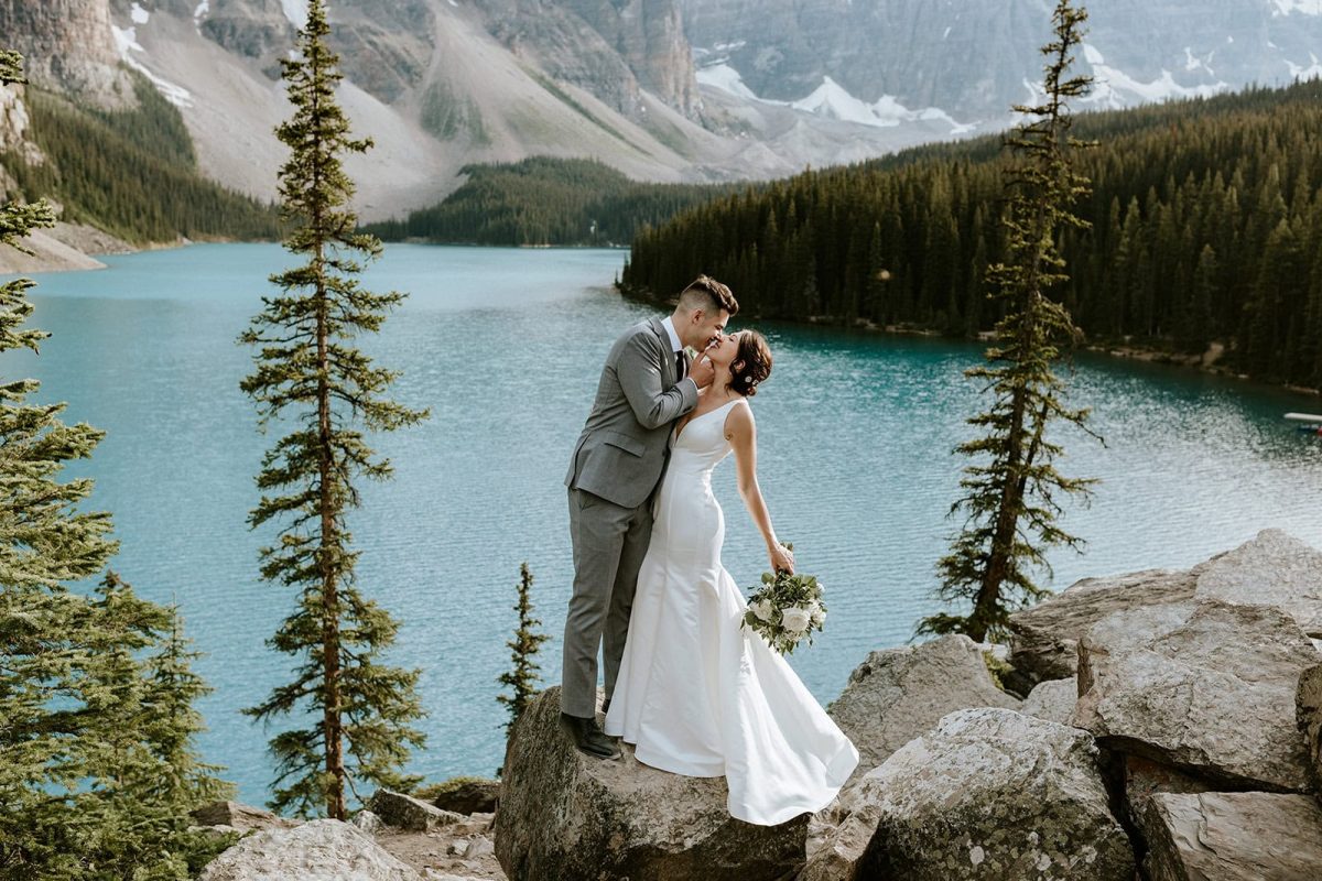 A bride and groom share a kiss on a rocky outcrop overlooking a turquoise lake and pine-covered mountains, capturing an intimate elopement moment. The photographer frames the bride’s bouquet, white dress, and groom’s gray suit against the stunning view.