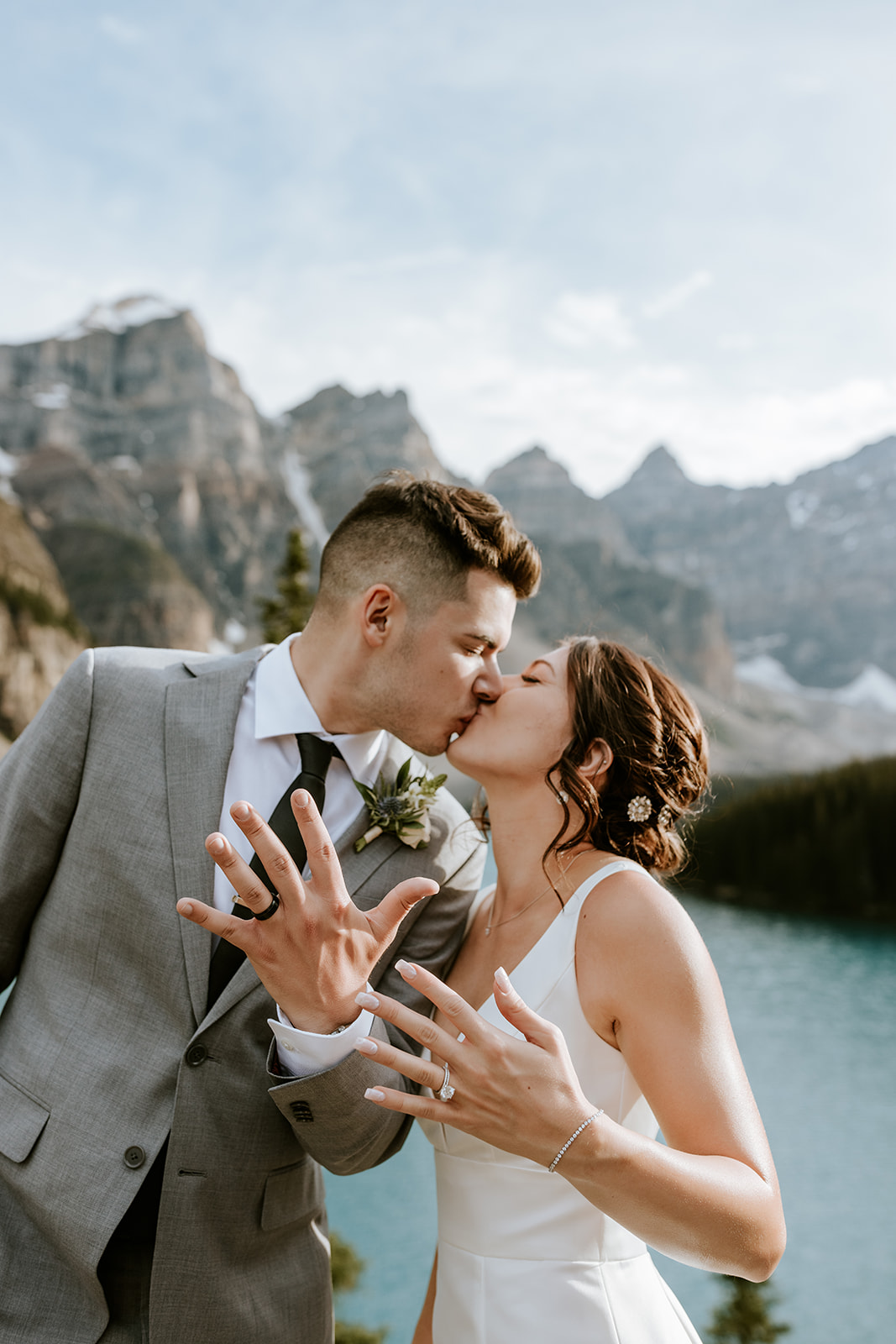 A newlywed couple shares a kiss by a mountain lake near Canmore, showing off their wedding rings. The groom wears a gray suit and the bride a white dress, framed by pine trees and rocky peaks in this intimate elopement moment.