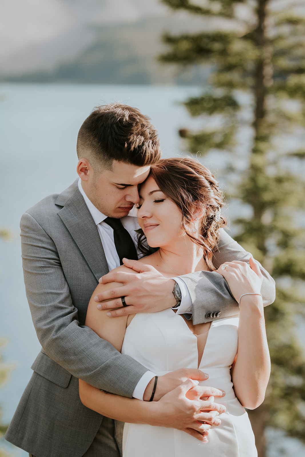A couple embraces lovingly outdoors near a lake in Calgary; the man in a grey suit hugs the woman in a white dress from behind. Captured by a photographer, they stand among trees and mountains, looking peaceful and content on their wedding day.